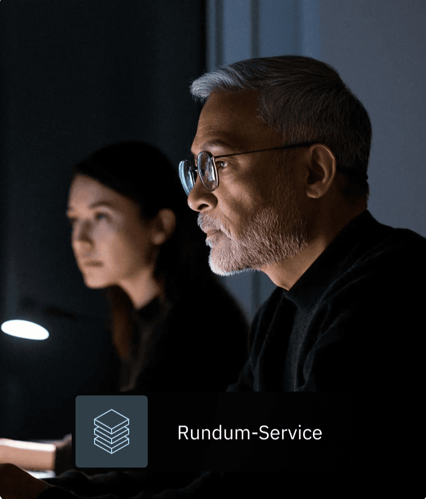 Mann mit Brille und Frau arbeiten in einem abgedunkelten Raum konzentriert am Bildschirm, eingeblendet der Hinweis „Rundum-Service".