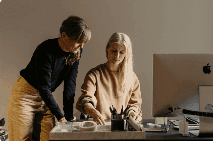 Zwei Frauen an einem Schreibtisch mit Computer betrachten gemeinsam Unterlagen.