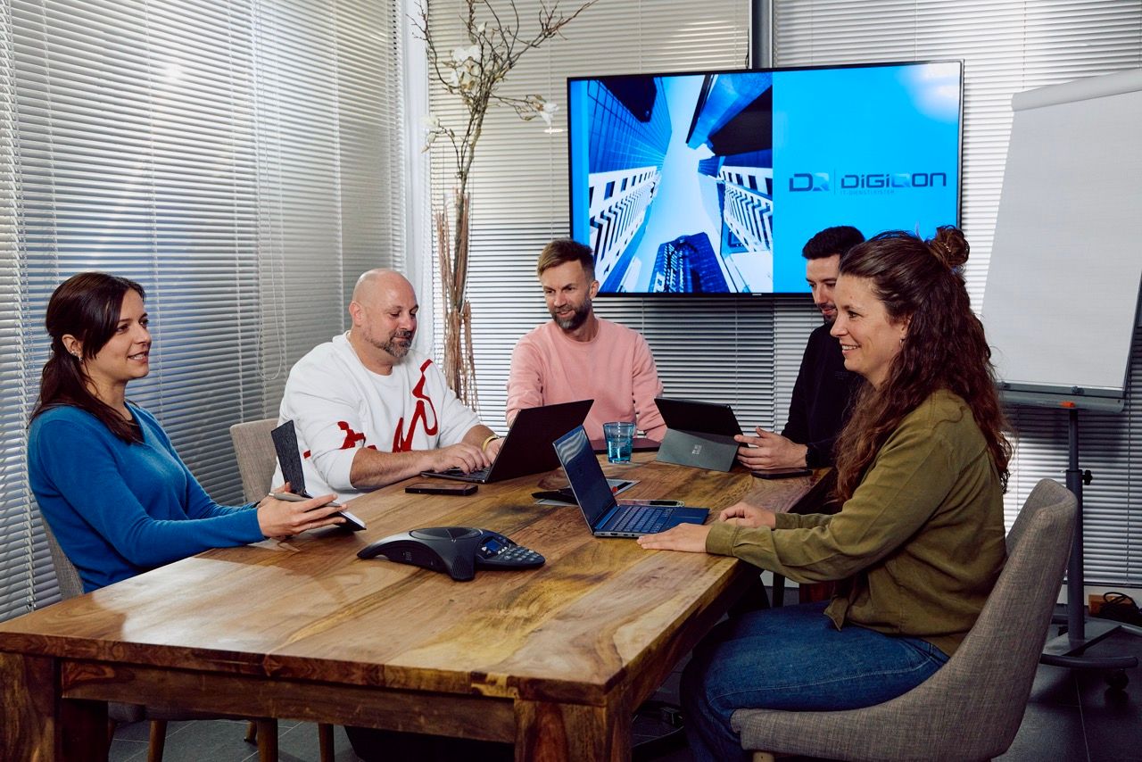Fünf Personen mit Laptops in einer Besprechung am Holztisch, im Hintergrund ein Bildschirm mit dem DigiQon-Logo.