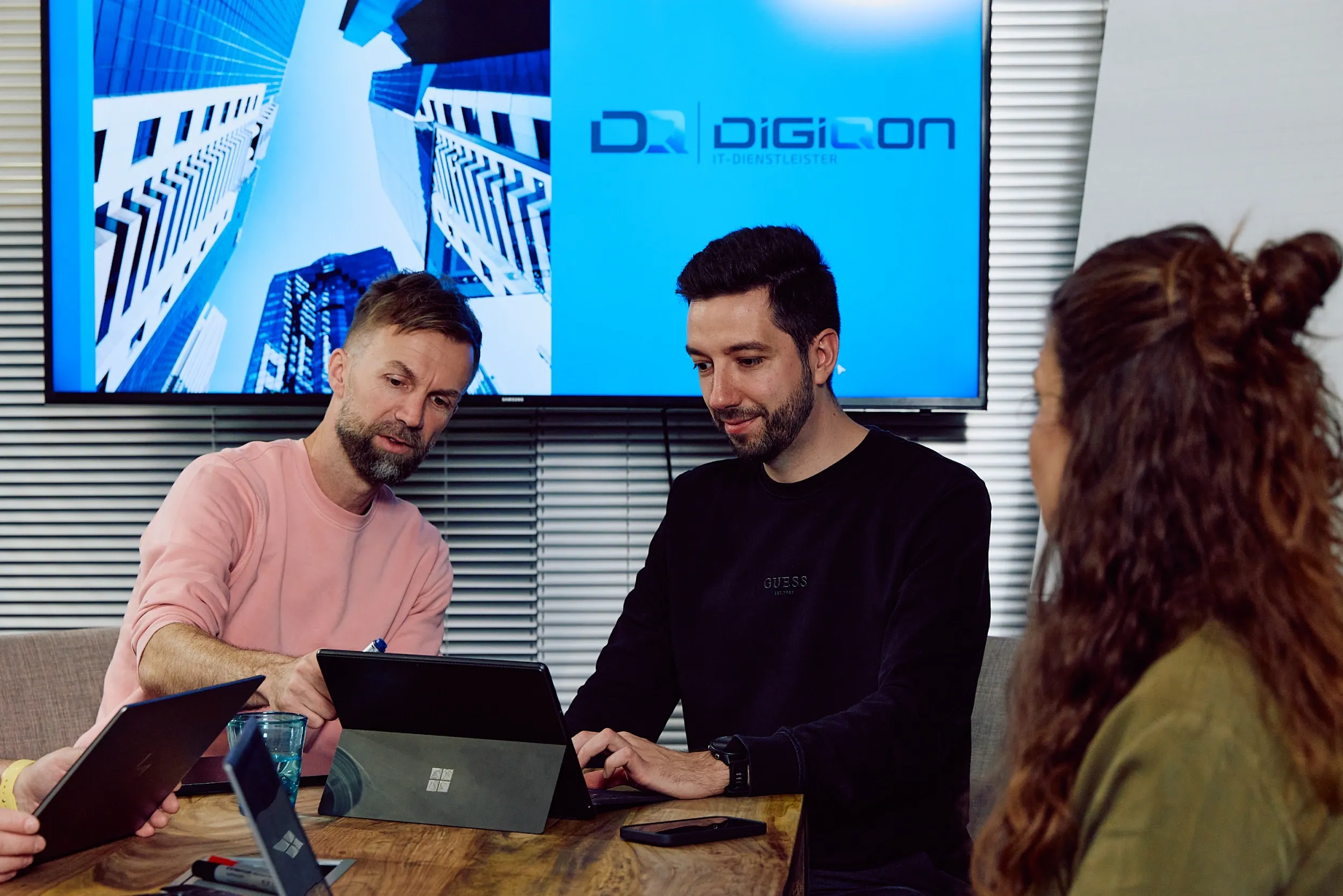 Drei Personen am Besprechungstisch mit Laptop, im Hintergrund ein Bildschirm mit dem DigiQon-Logo.