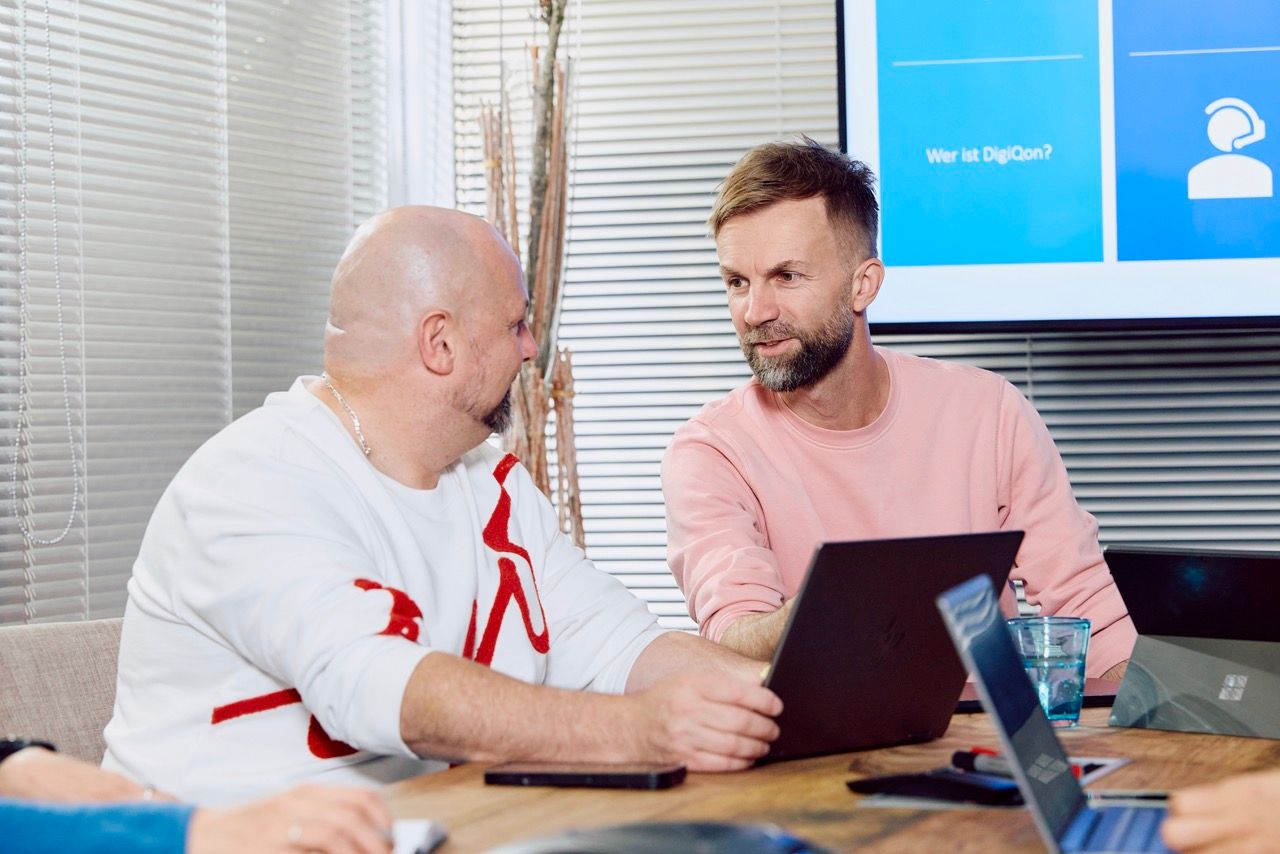 Zwei Männer im Gespräch an einem Tisch mit Laptops, im Hintergrund eine Präsentation mit der Frage „Wer ist DigiQon?“.