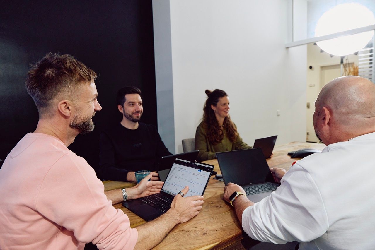Vier Personen sitzen mit Laptops um einen langen Holztisch in einem hellen Besprechungsraum.