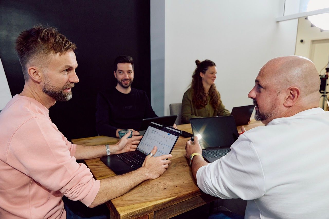 Vier Personen sitzen mit Laptops an einem Holztisch und besprechen sich in einem Meeting.