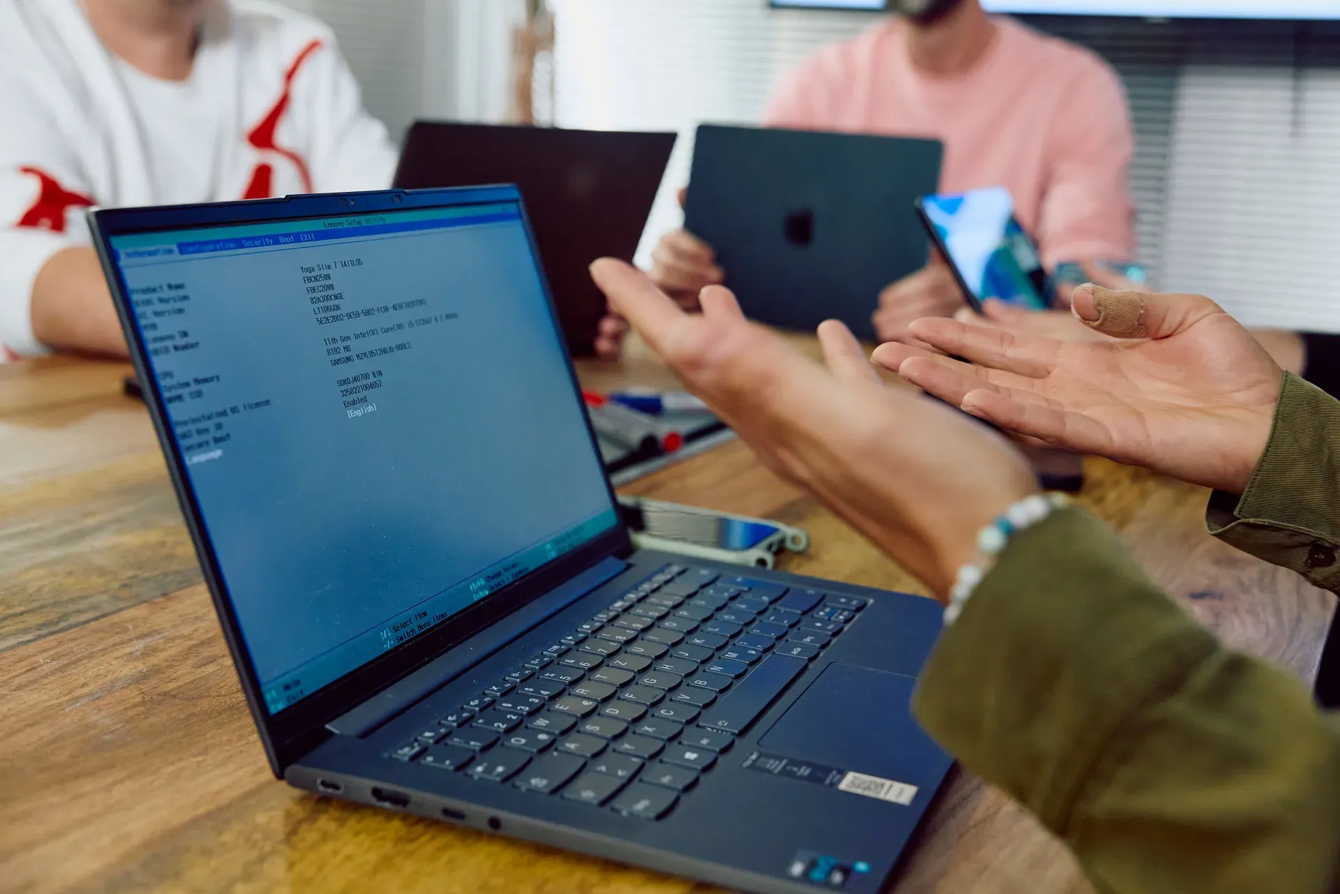 Aufgeklappter Laptop mit BIOS-Setup auf dem Bildschirm, im Hintergrund Personen mit weiteren Geräten am Tisch.