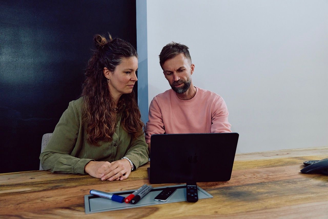 Frau und Mann sitzen an einem Holztisch im Besprechungsraum und blicken gemeinsam auf einen Laptop.