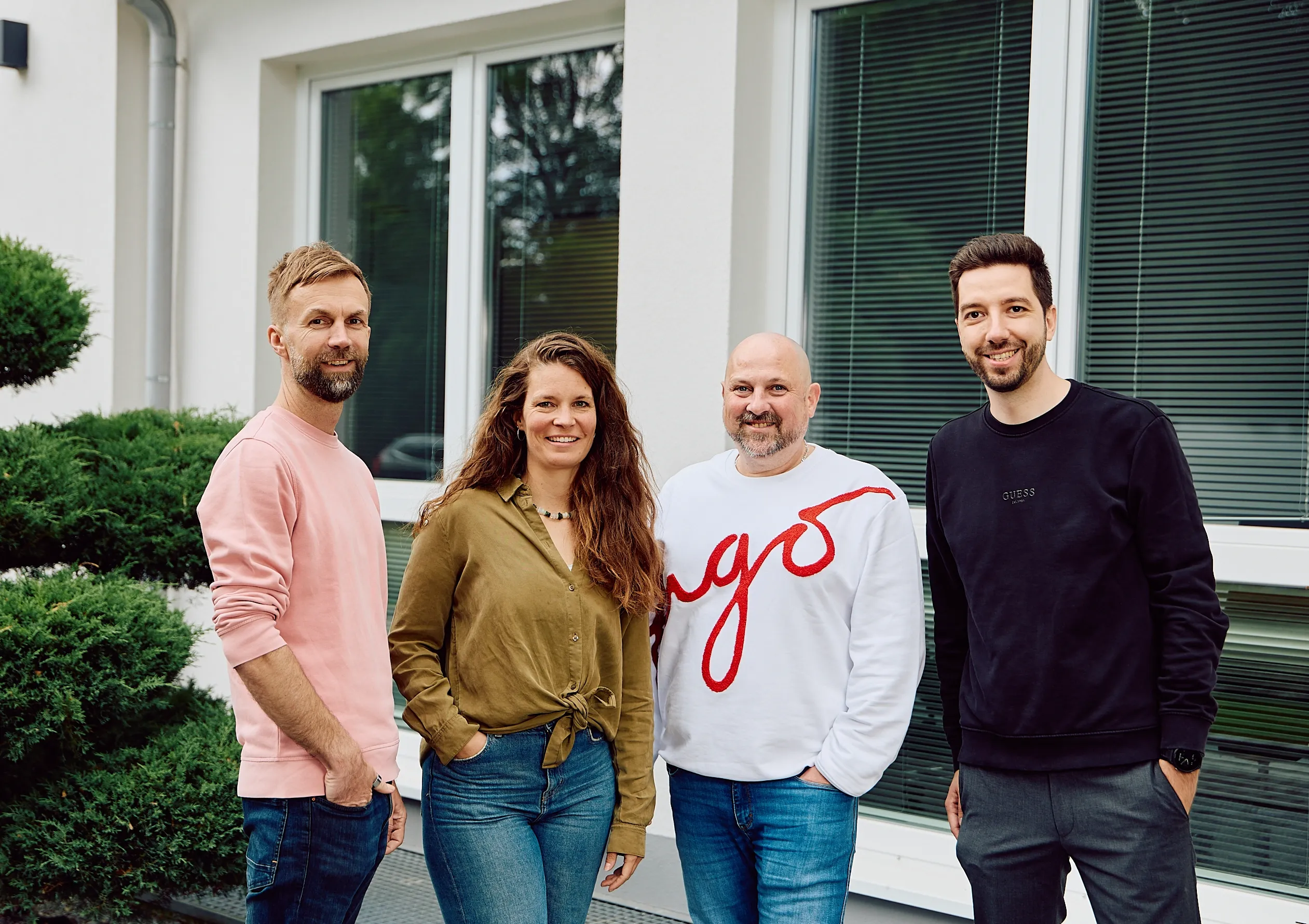 Das DigiQon-Team vor dem Büro in Grünwald: vier Mitarbeitende lächeln gemeinsam vor der hellen Eingangsfront.