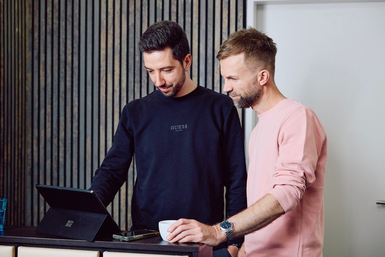 Zwei Männer stehen an einem Sideboard und schauen gemeinsam auf ein Tablet, einer hält eine Kaffeetasse.