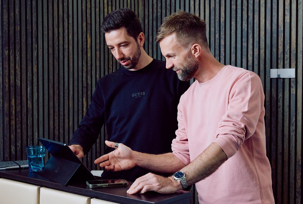 Zwei DigiQon-Mitarbeiter besprechen gemeinsam ein IT-Konzept an einem Tablet vor einer dunklen Holzlamellen-Wand.
