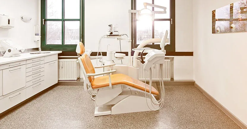 Zahnarztpraxis Hummel: Behandlungszimmer mit einem orangefarbenen Behandlungsstuhl an zwei Fenstern.
