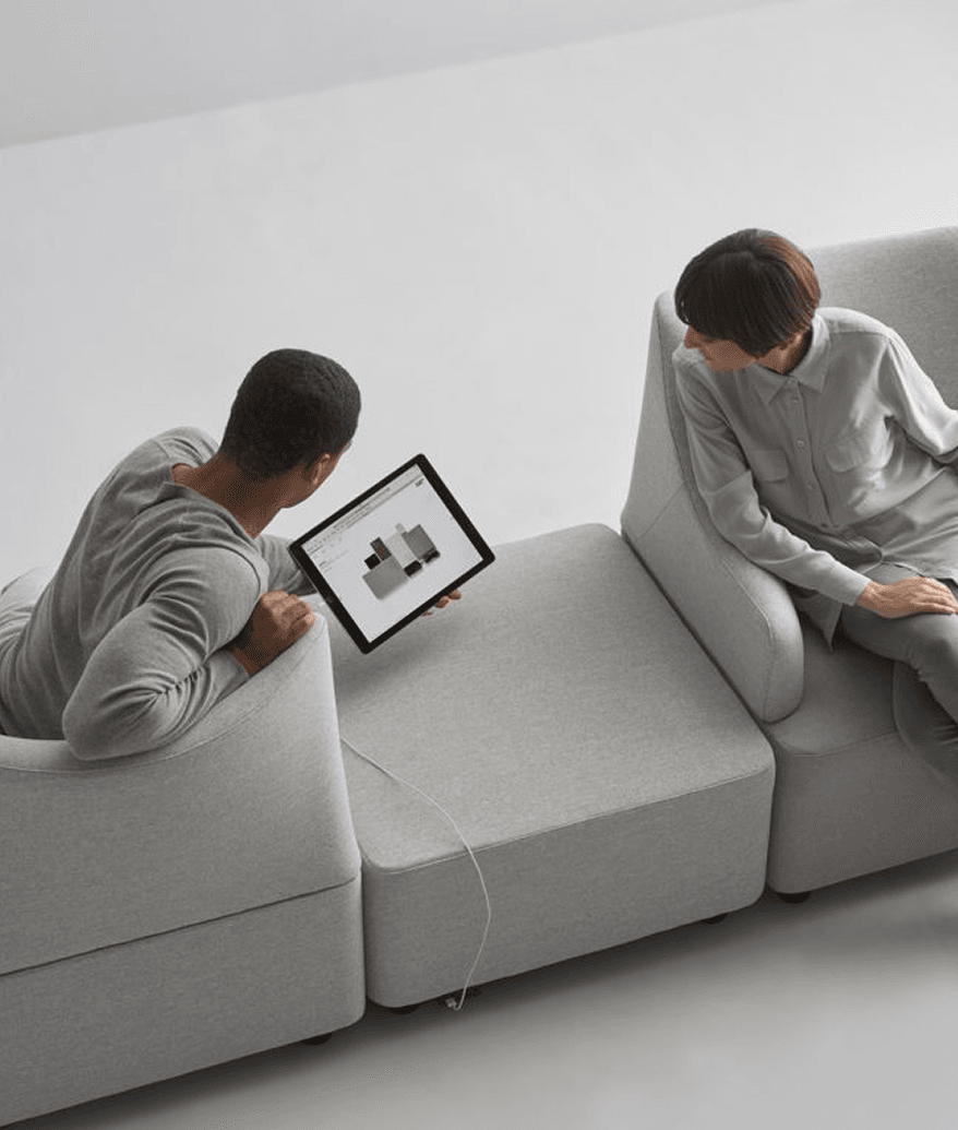 Zwei Personen sitzen auf hellgrauen Sofamodulen, eine zeigt der anderen ein 3D-Modell auf einem Tablet.