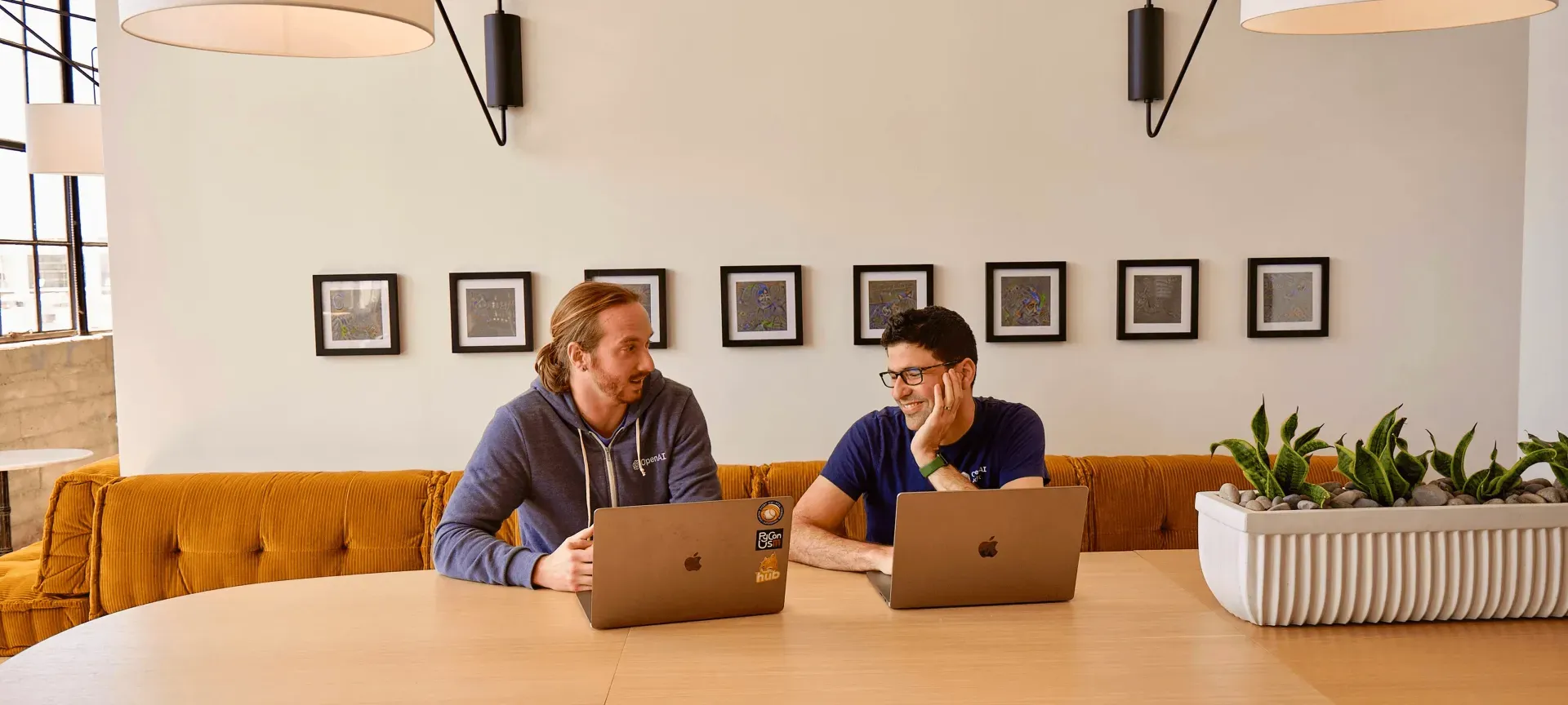 Zwei Männer mit Laptops sitzen an einem Tisch vor einer orangefarbenen Sitzbank in einem hellen Büro.